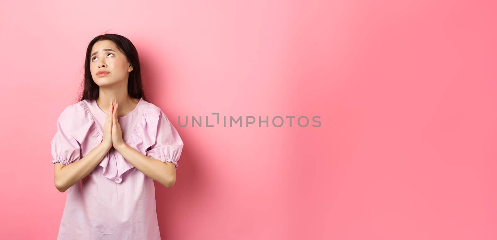 Hopeful asian girl praying god, looking up to plead or pray, making wish, supplicating with dramatic face, standing against pink background.