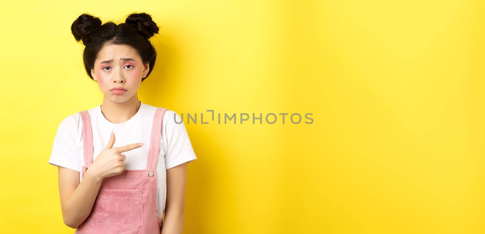 Sad and disappointed asian girl frowning, feel unfair, pointing finger right at bad thing, complaining on something upsetting, yellow background.