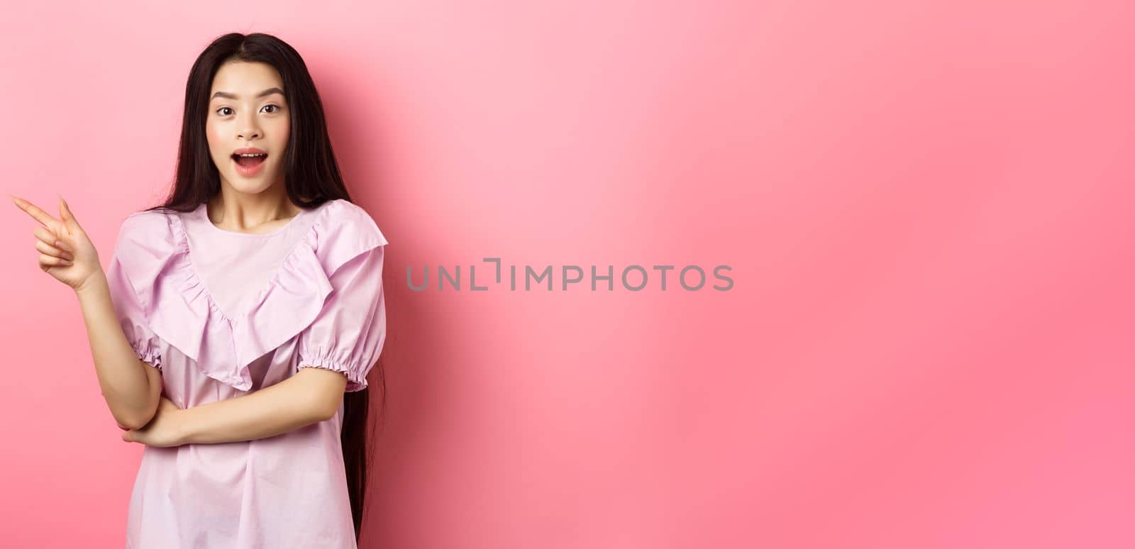 Excited beautiful asian teen girl pointing finger left, open mouth amazed and looking at camera, showing cool promo deal, pink background.