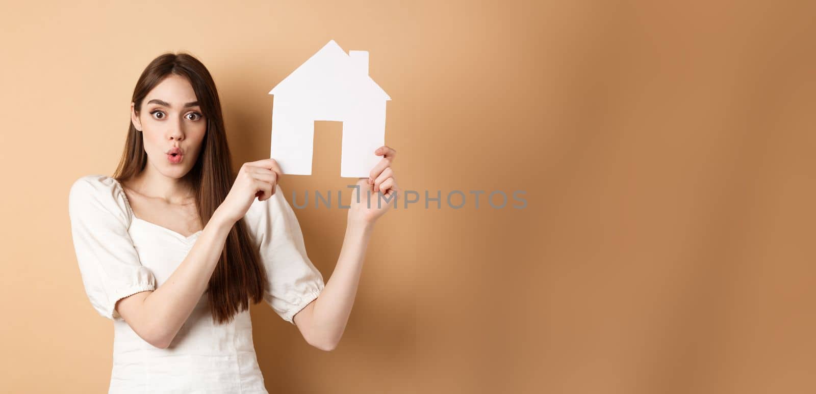 Real estate. Excited woman showing paper house cutout and saying wow with amazed face, buying property, standing on beige background by Benzoix