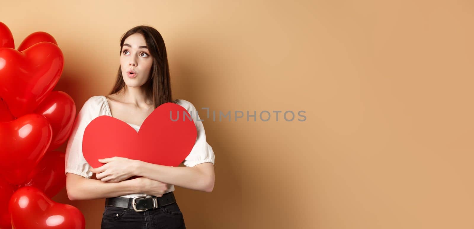 Valentines day and love concept. Intrigued tender girl hugging big red heart cutout and stand near balloons, looking aside at logo, saying wow, beige background.