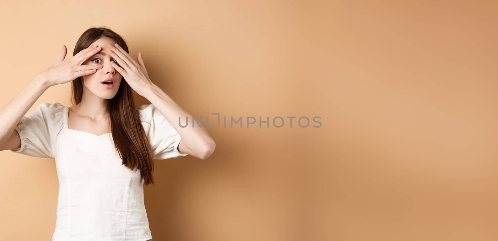 Curious girl peeking through fingers at something interesting, cannot wait to see surprise, look intrigued at camera, beige background by Benzoix