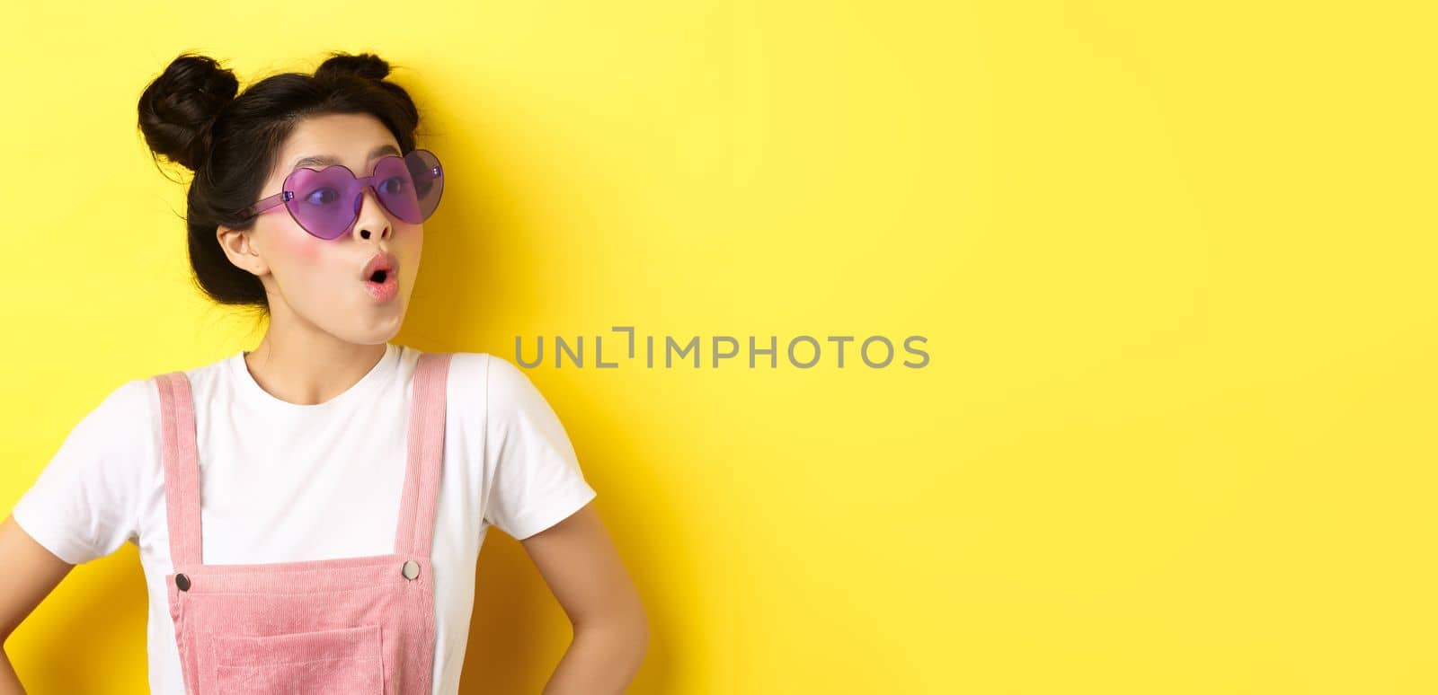 Summer and fashion concept. Close-up of surprised asian teen girl in heart-shaped sunglasses looking right side with excited face, standing on yellow background.
