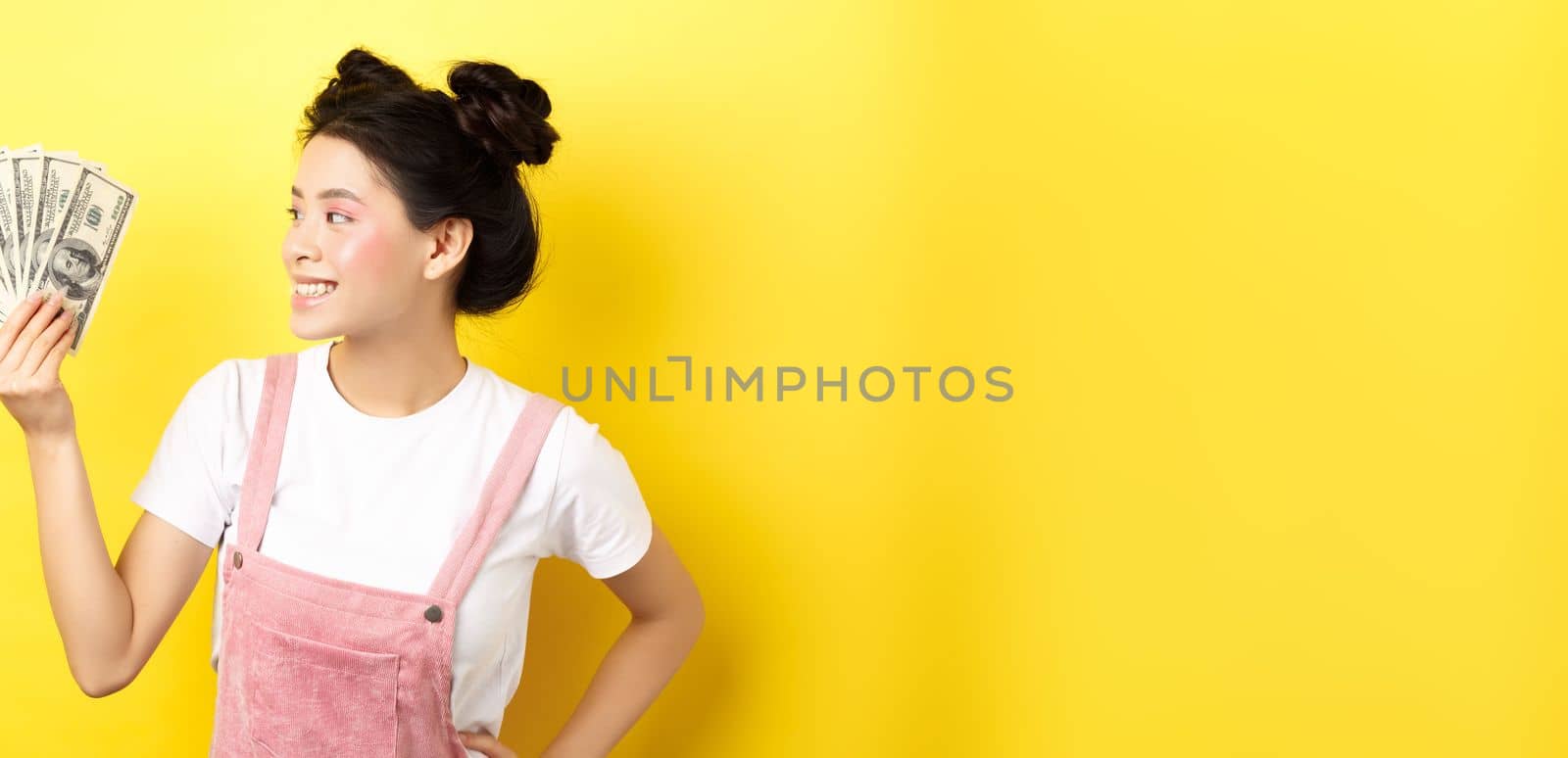 Shopping. Rich and stylish asian female model showing money, looking at dollar bills with pleased smile, standing on yellow background by Benzoix
