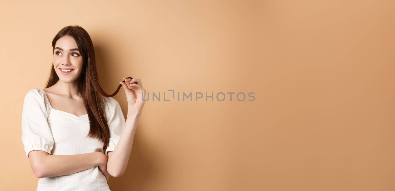 Romantic pensive girl thinking, looking left at empty space and playing with hair strand, smiling dreamy, standing in white dress on beige background.