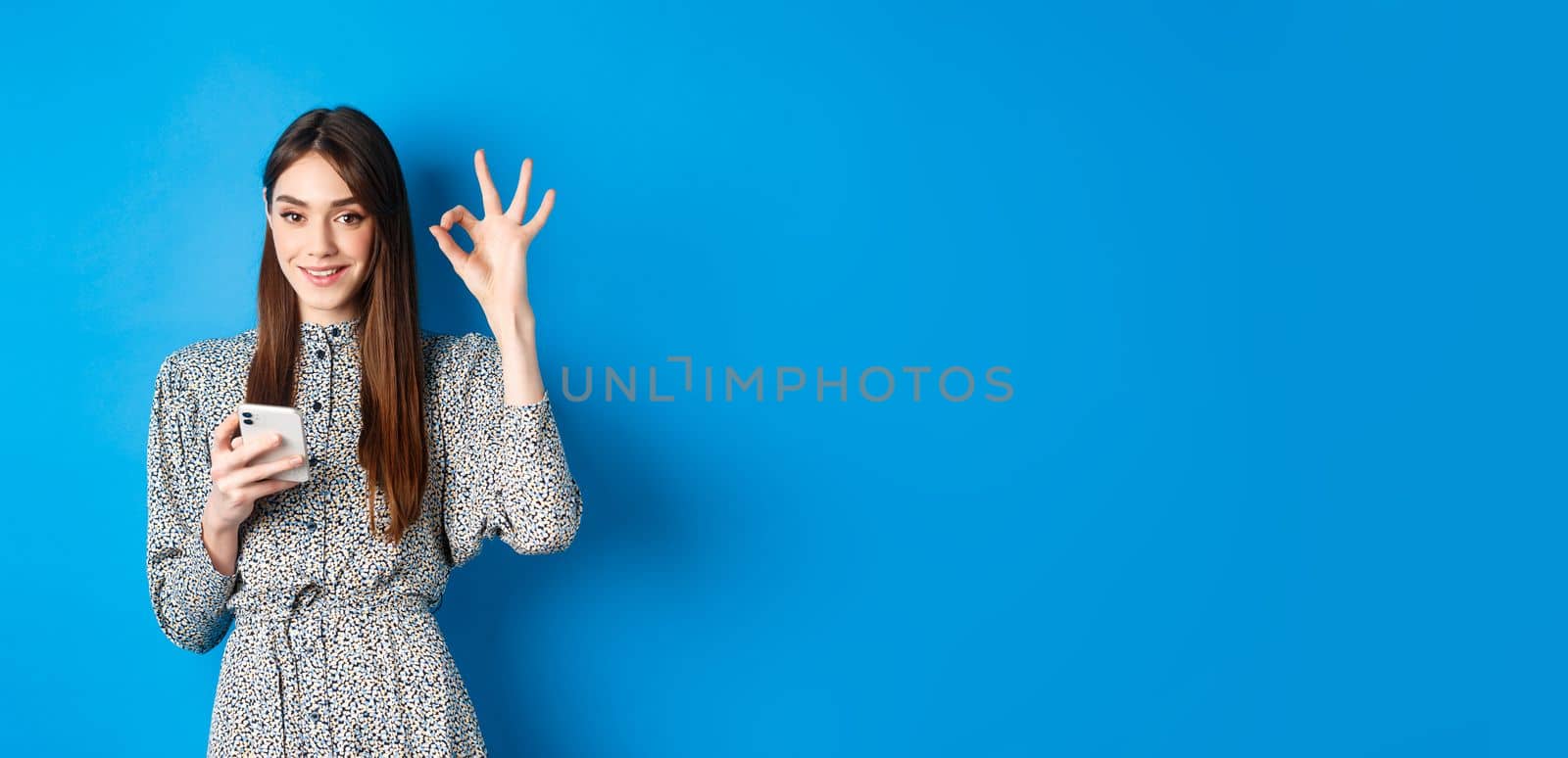 Beautiful young woman in dress showing okay sign and using smartphone, smiling at camera, standing on blue background.