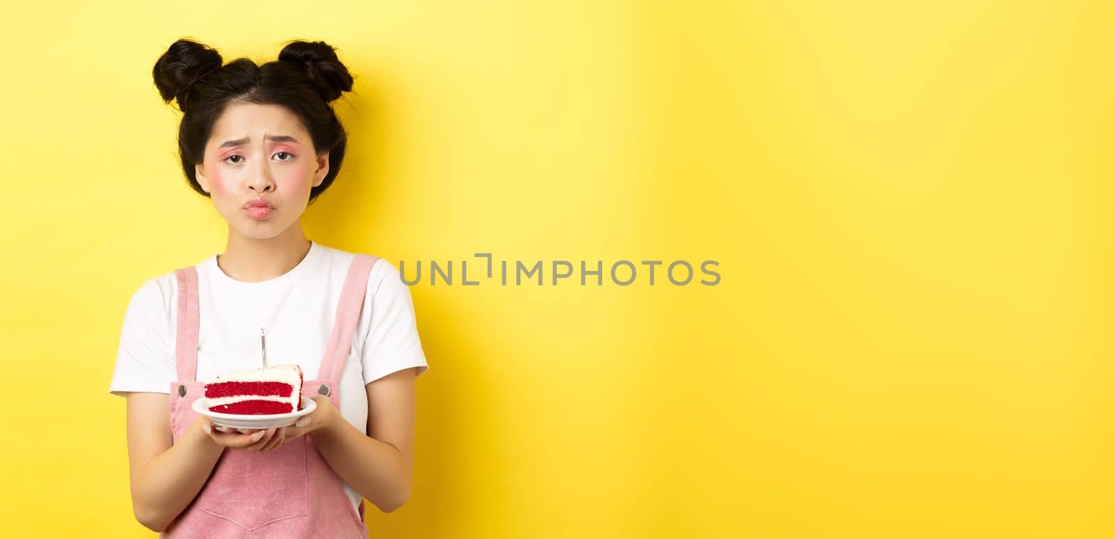 Sad and lonely birthday girl frowning upset, holding birthday cake with candle, making wish, standing on yellow background by Benzoix