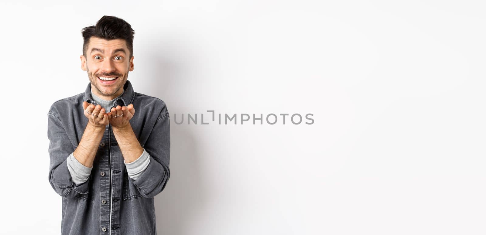 Lovely young man sending air kiss at camera and smiling. Hopeless romantic showing his love, holding something in hands, standing on white background.
