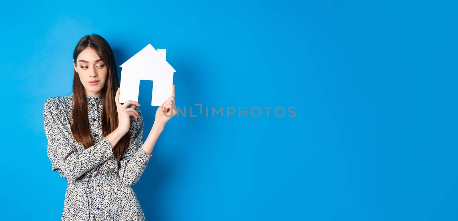 Real estate, realtors and insurance concept. Beautiful young woman in dress holding paper house cutout, looking aside dreamy, searching apartment for rent, blue background.