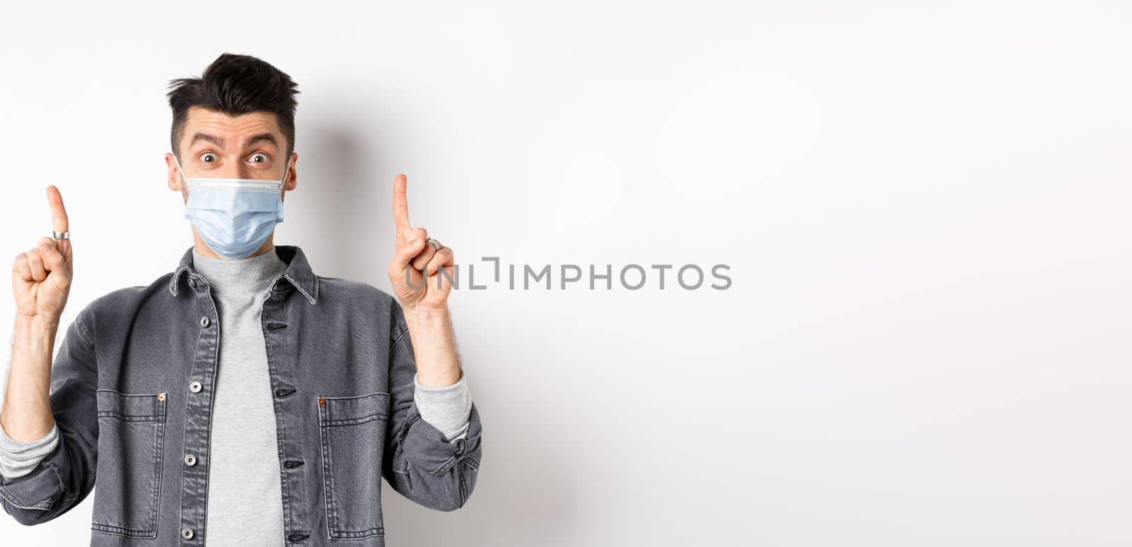 Pandemic lifestyle, healthcare and medicine concept. Excited man advertising in medical mask, pointing fingers up and look amazed, standing on white background.