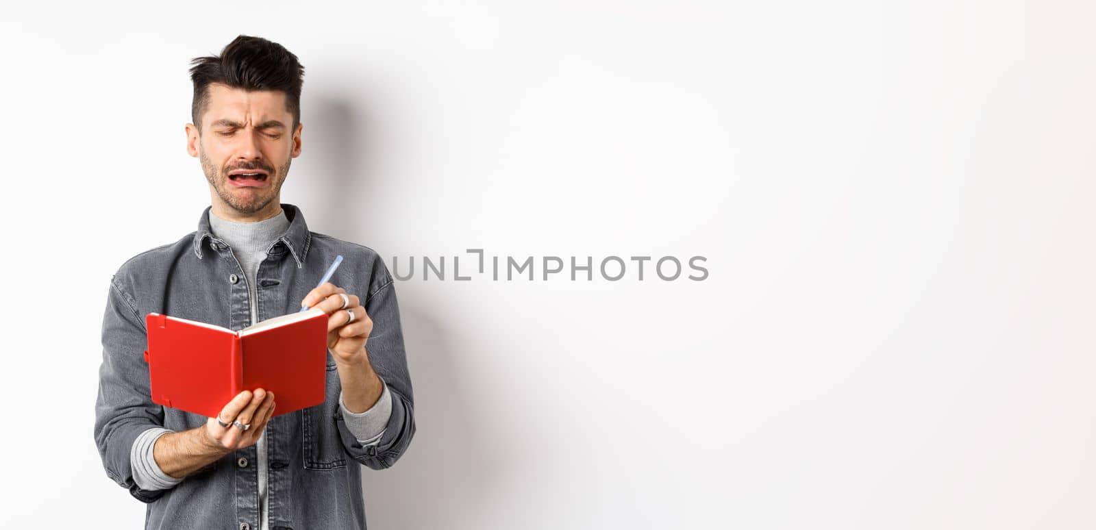 Sad crying guy writing in his diary with miserable face, look distressed while making notes in journal or planner, standing against white background.