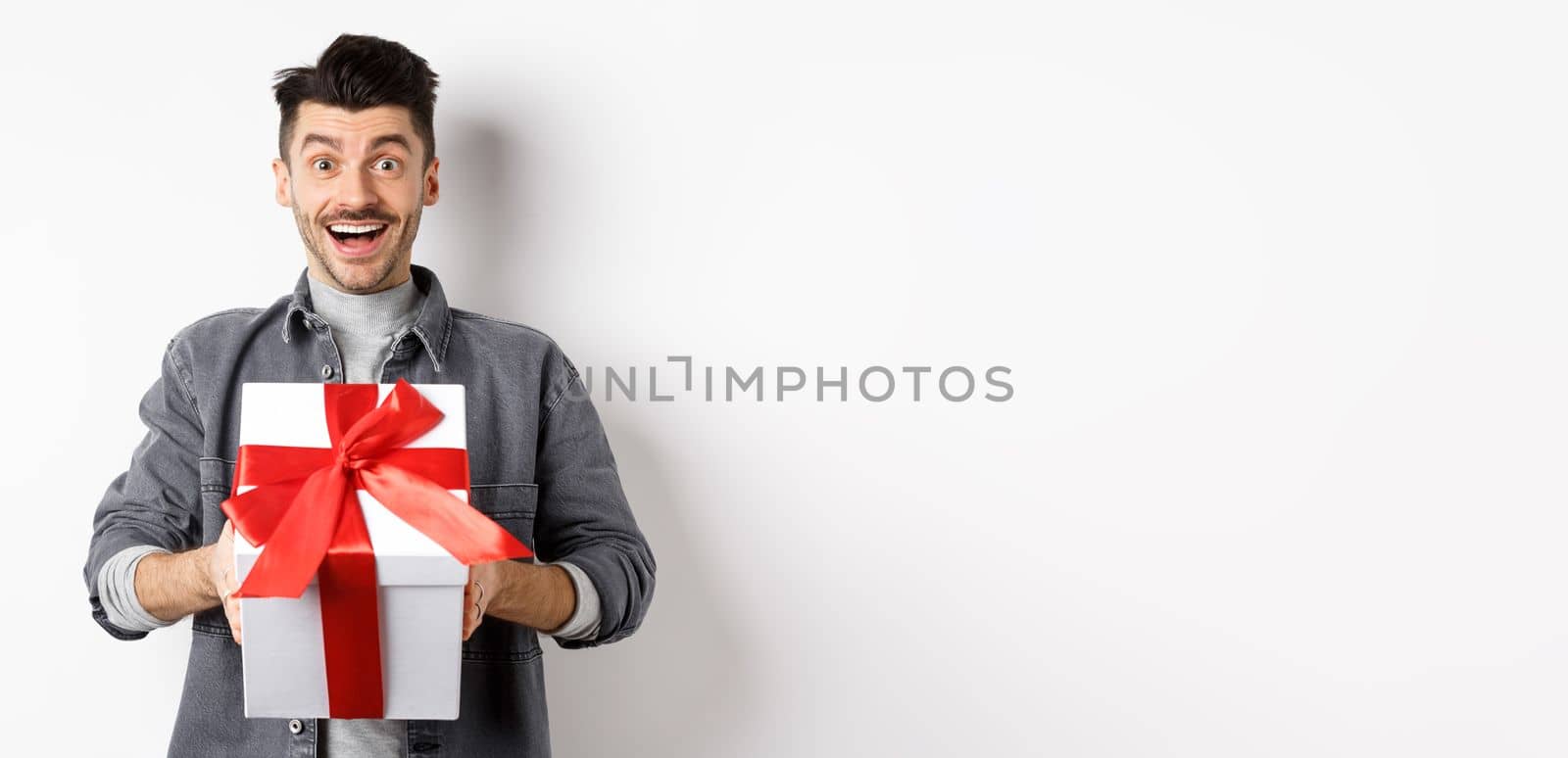 Surprised young man smiling excited, holding big gift box on valentines day holiday, receive surprise present, standing amazed on white background.