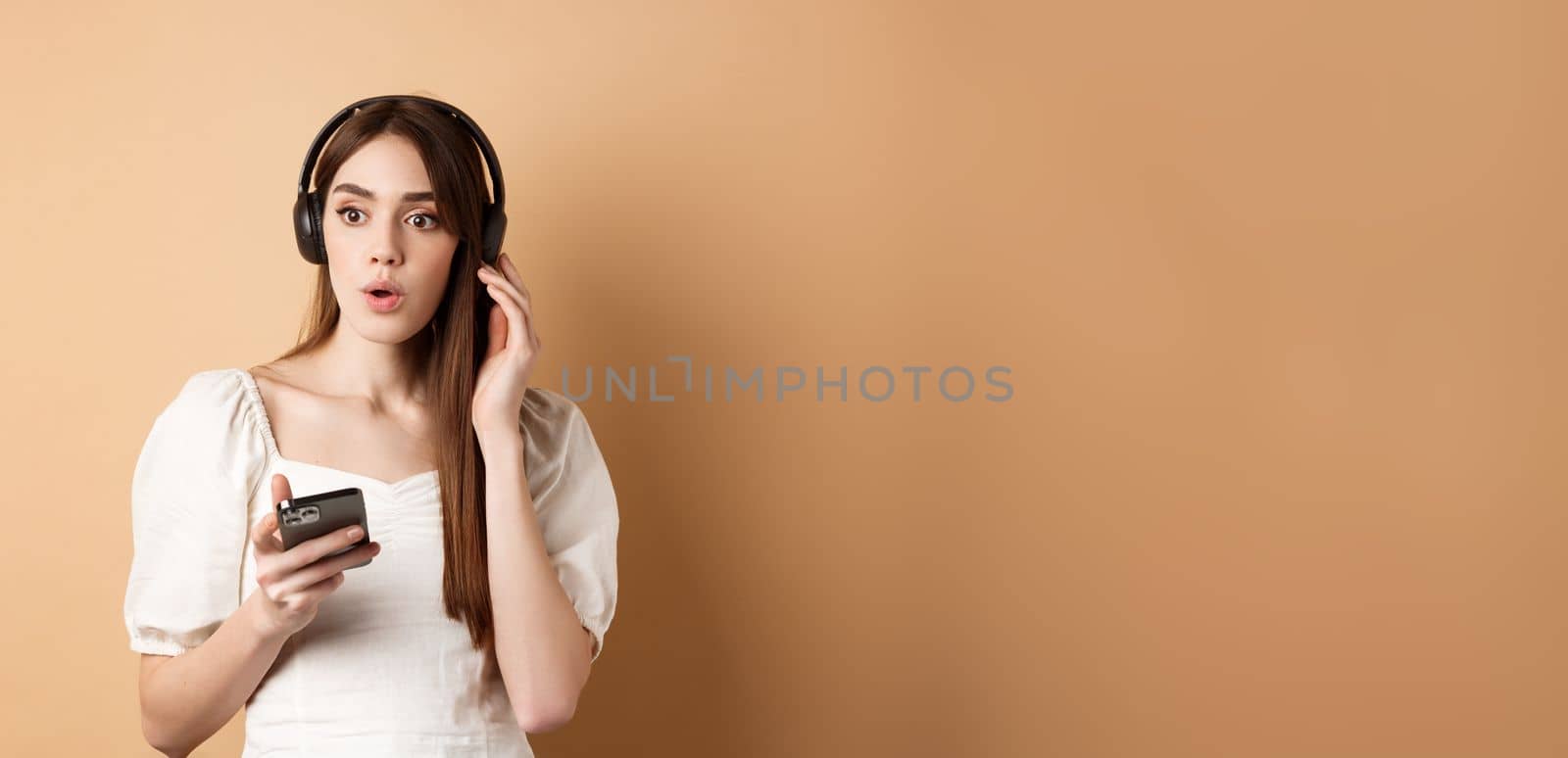 Wow awesome tunes. Excited young girl amazed with new song, listening music in headphones and look impressed, holding cell phone, beige background by Benzoix