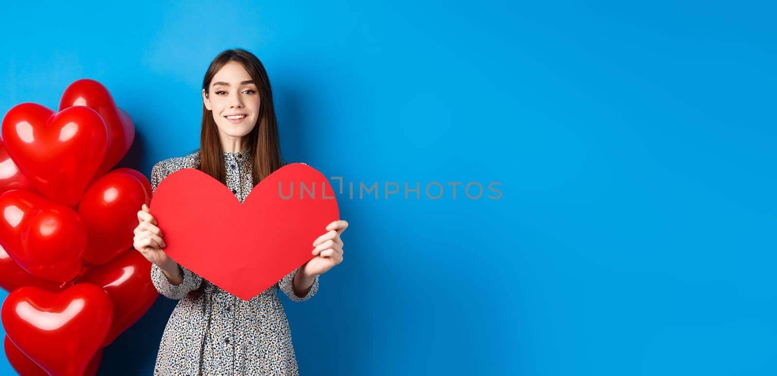 Valentines day. Lovely young woman in dress celebrating lovers holiday, showing valentine card and smiling, standing near red heart balloons on blue background by Benzoix