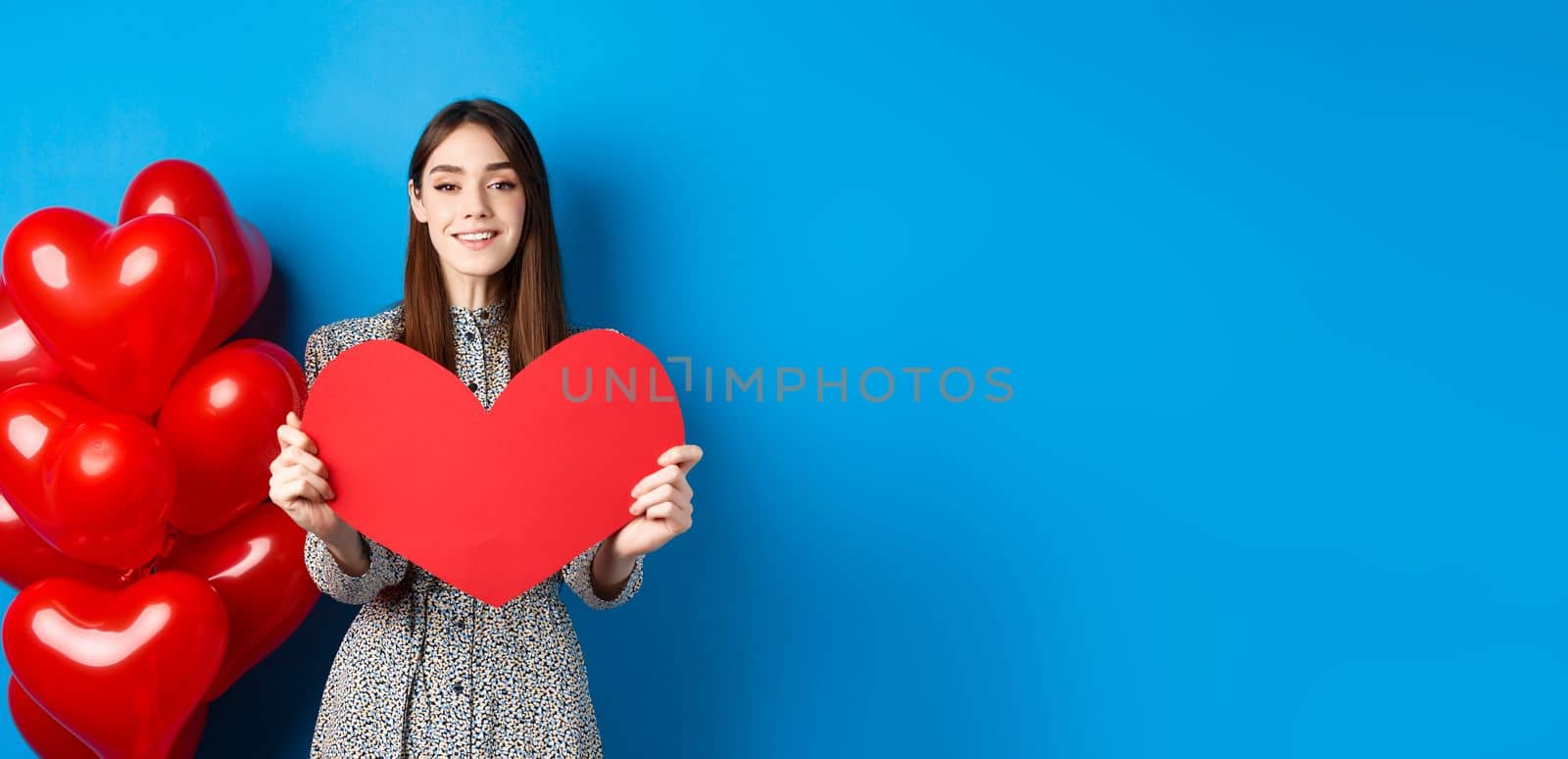 Valentines day. Lovely young woman in dress celebrating lovers holiday, showing valentine card and smiling, standing near red heart balloons on blue background.