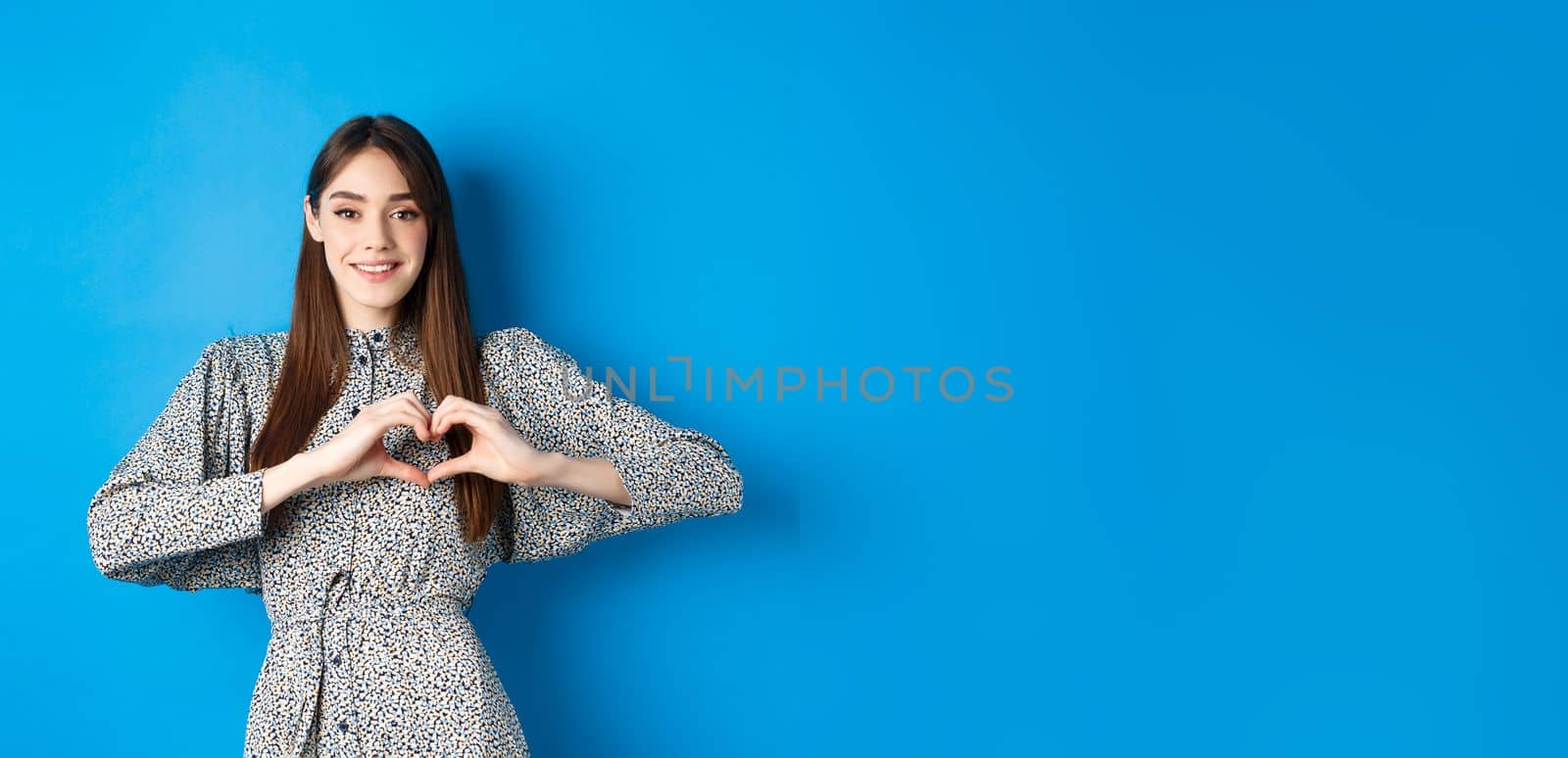 Pretty girl in romantic dress showing I love you heart gesture, smiling at camera, express sympathy and romance, standing on blue background.
