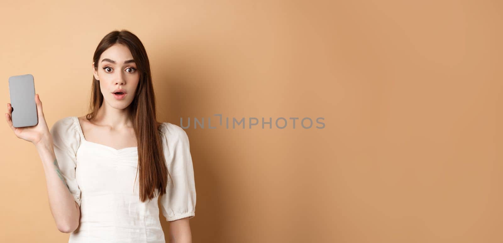 Excited pretty girl showing empty cell phone screen, look amazed at camera, standing on beige background.