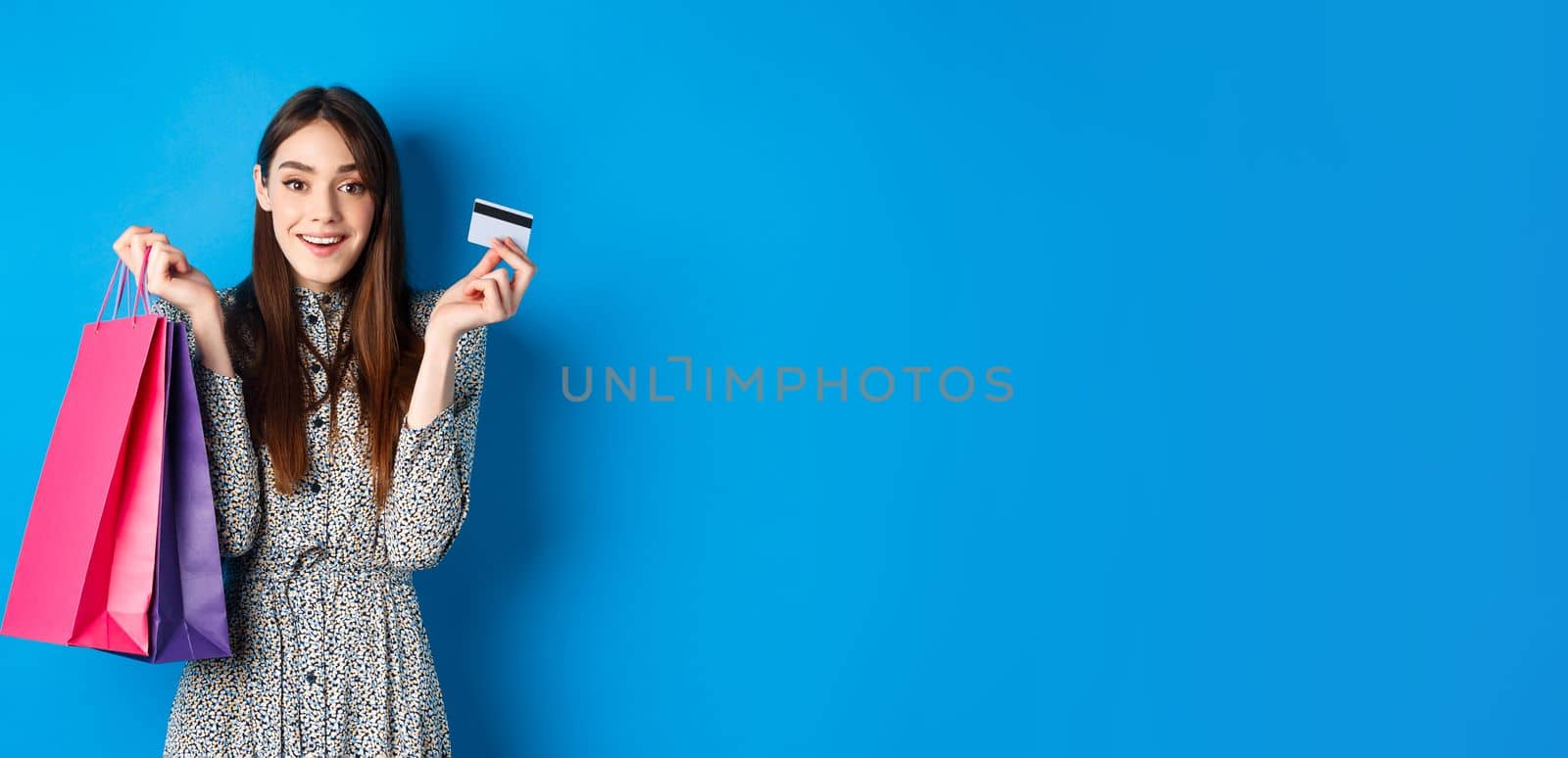 Excited beautiful female model shopping with plastic credit card, holding shop bags and smiling happy at camera, blue background.