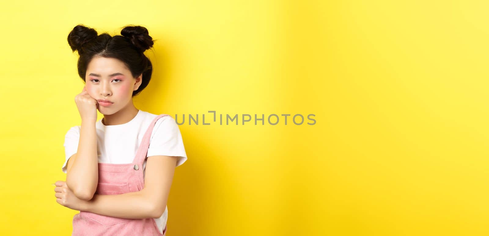 Bored asian teen girl with stylish makeup and summer clothes, looking reluctant and indifferent, standing on yellow background.