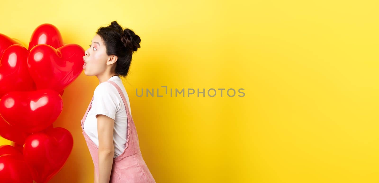 Valentines day and relationship concept. Profile of young asian woman scream of surprise, say wow and looking left amazed, standing near red balloons, yellow background.