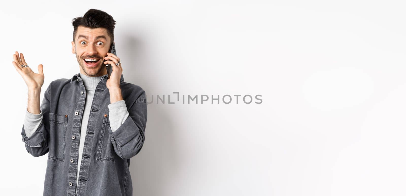 Excited man talking on cellphone and hear good news, rejoicing and raising hand with happy smile, standing against white background by Benzoix