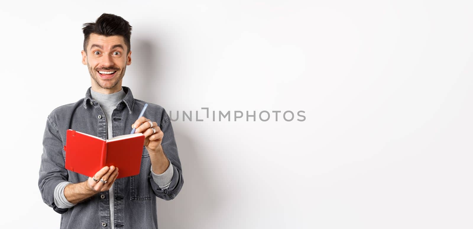 Creative young man smiling excited while taking notes, writing down ideas in planner, holding red journal and looking happy, standing against white background.
