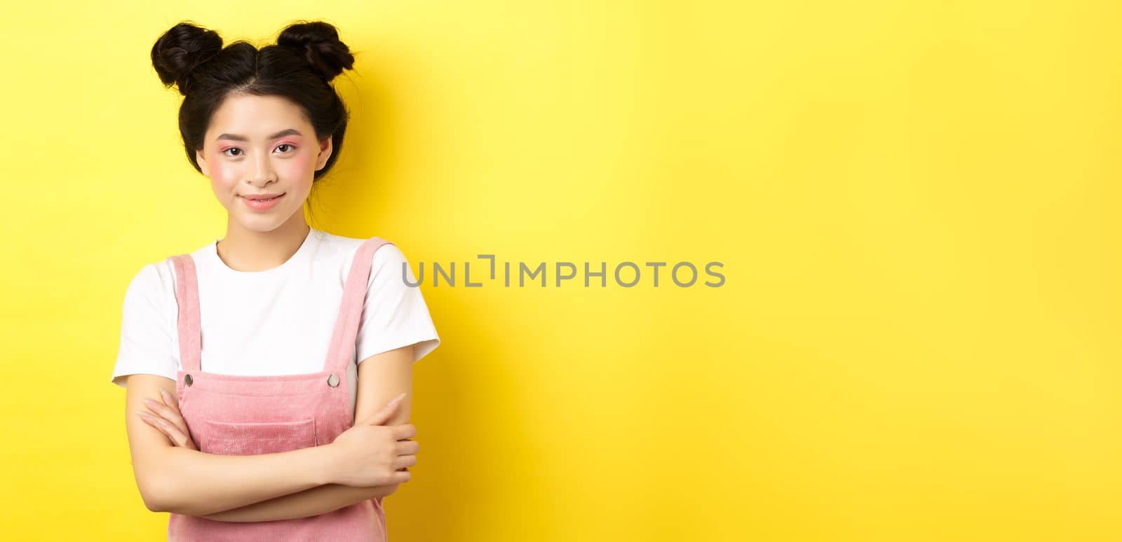 Beauty asian girl with glamour makeup, cross arms on chest and smiling tenderly at camera, standing on yellow background.