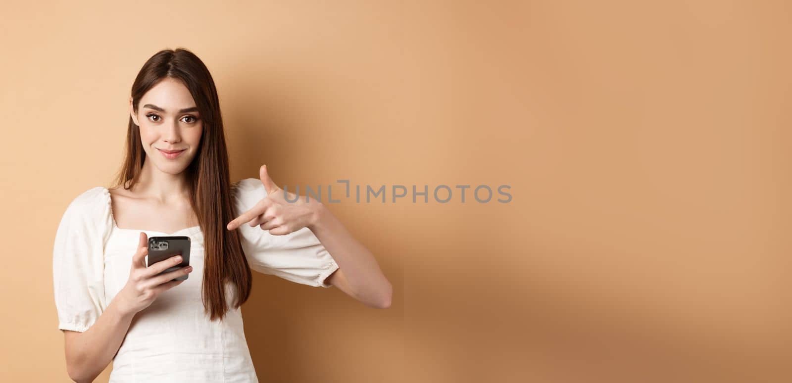 Attractive woman smiling and pointing at smartphone, showing online offer on phone, standing on beige background by Benzoix