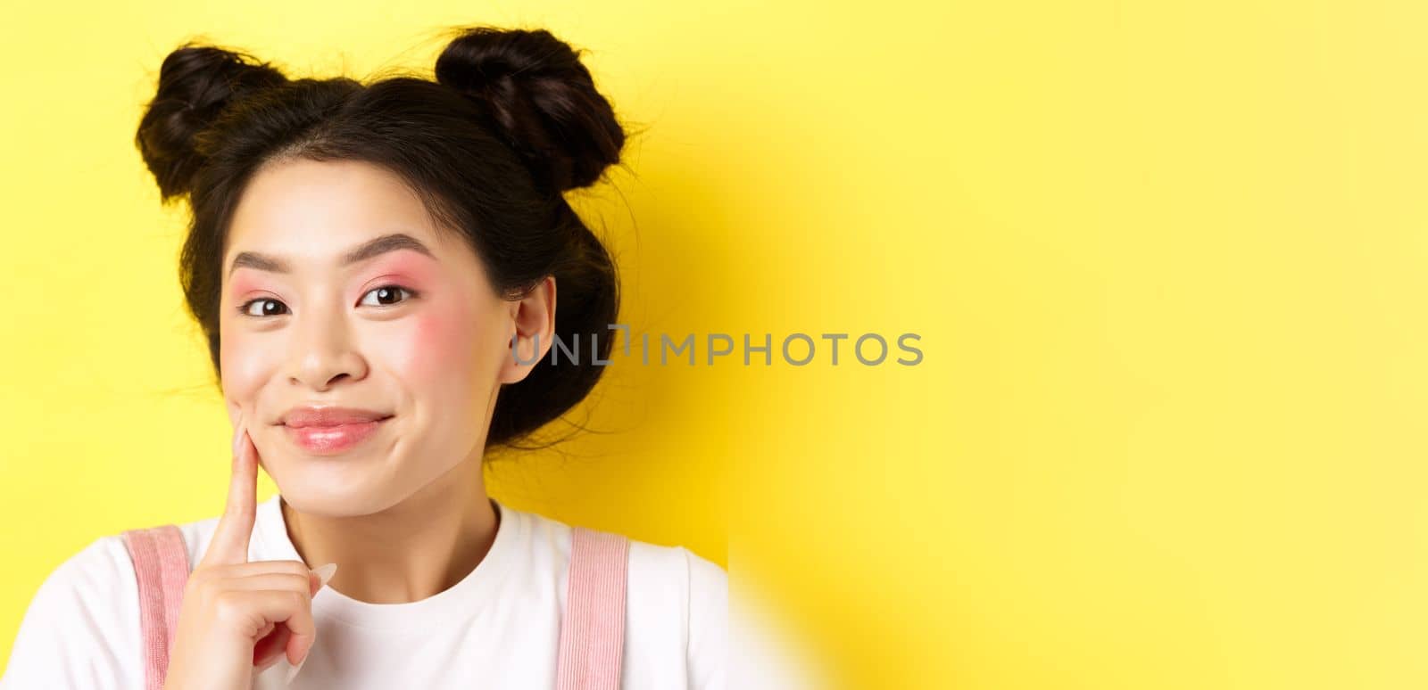Beauty. Stylish asian girl with bright glamour makeup, touching soft and shiny facial skin, smiling happy at camera, standing on yellow background by Benzoix