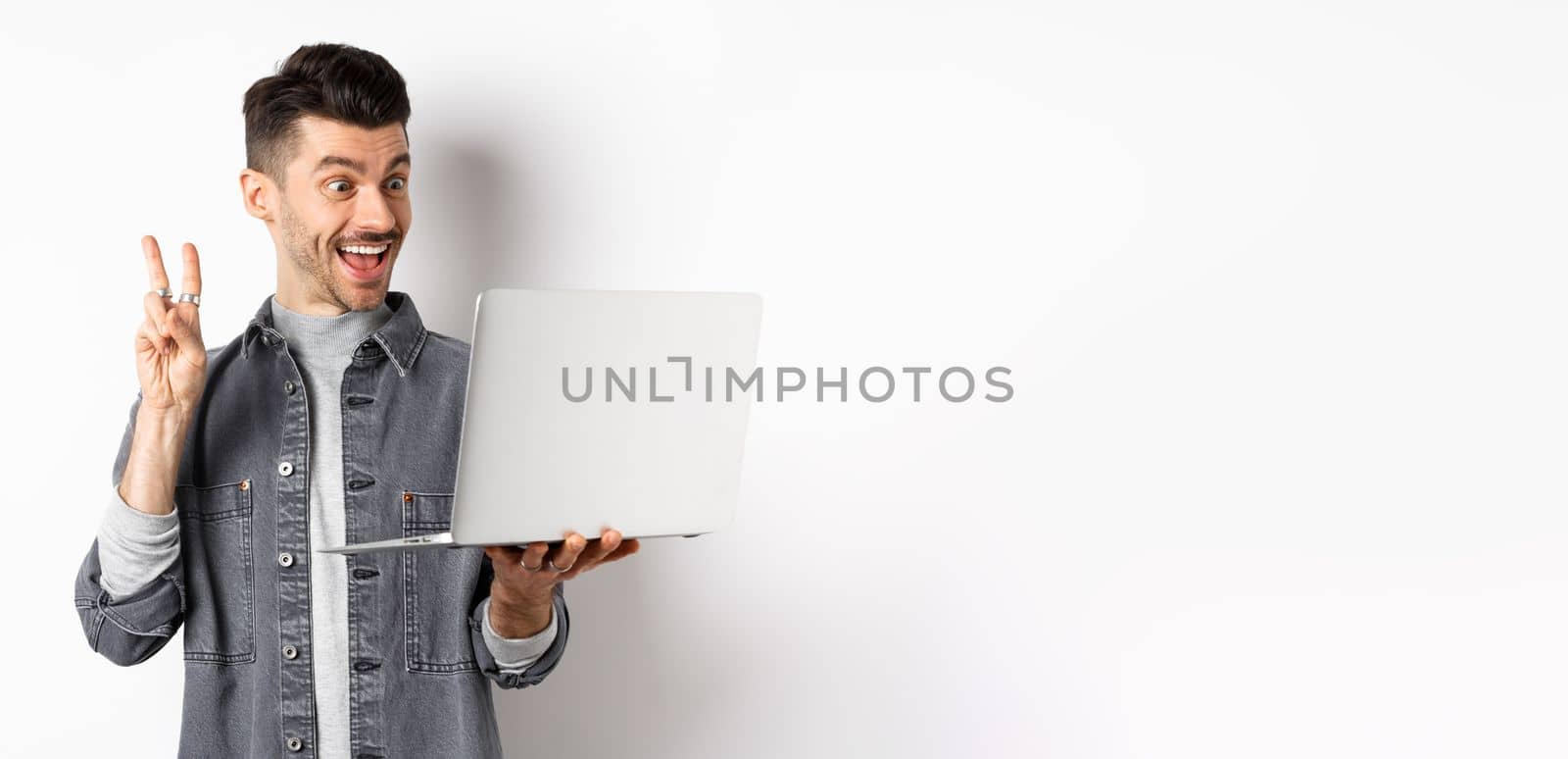 Funny young man video chat on laptop, showing v-sign and smiling at computer camera, standing against white background.