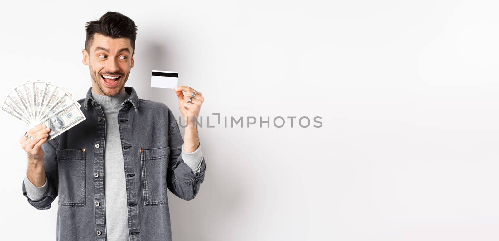 Excited guy holding plastic credit card and dollar bills, standing amused on white background.
