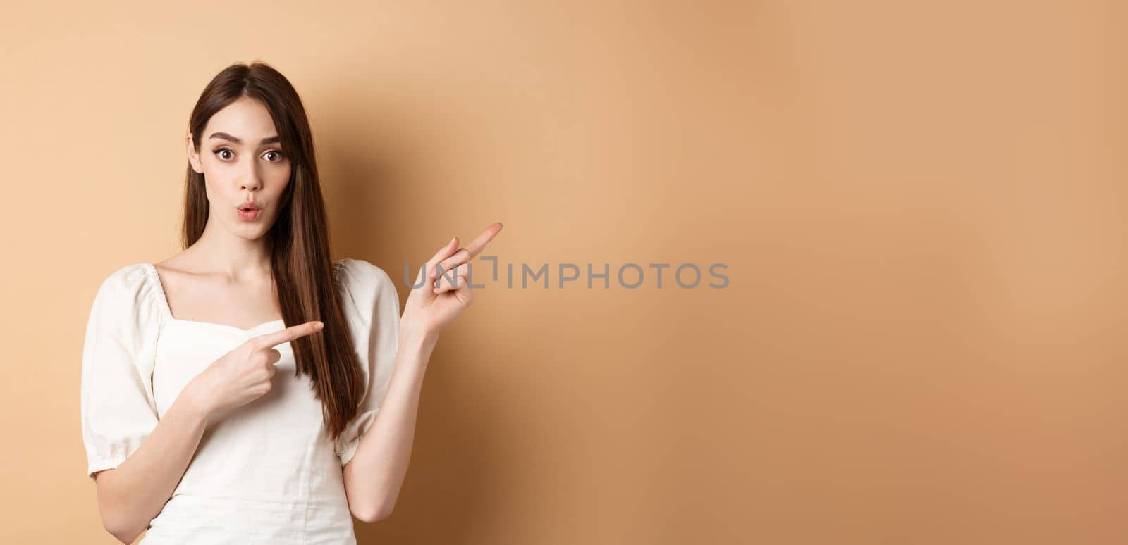 Wow look here. Impressed caucasian woman in white dress pointing fingers right and look curious, asking question about product, beige background.