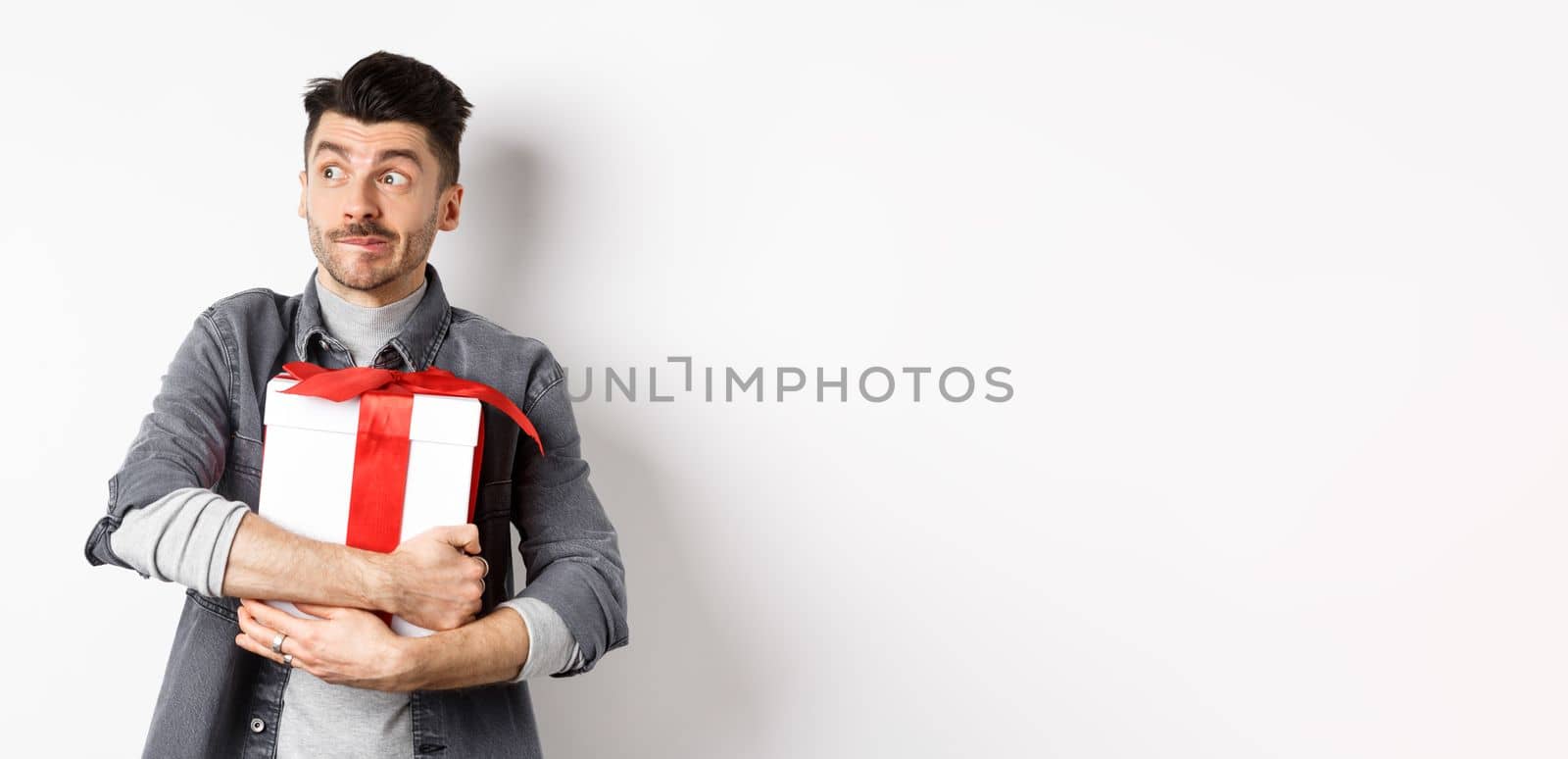 Excited man hugging romantic gift for girlfriend, waiting for valentines date with lover, looking at empty space logo with happy face, standing on white background.