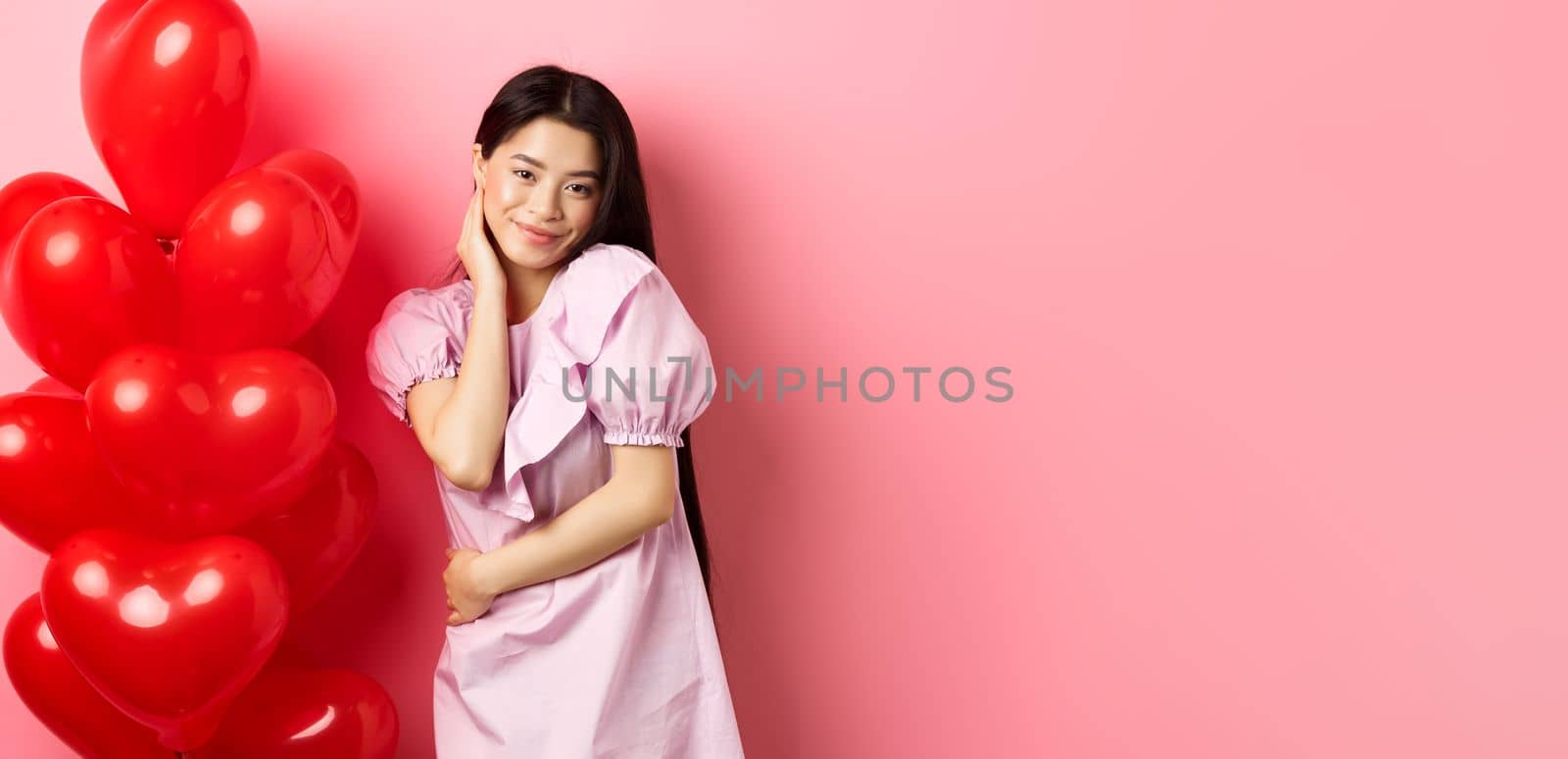 Beautiful asian girl in dress smiling coquettish, flirting on valentines day, looking sensual at camera, posing near valentines heart balloons, pink background.