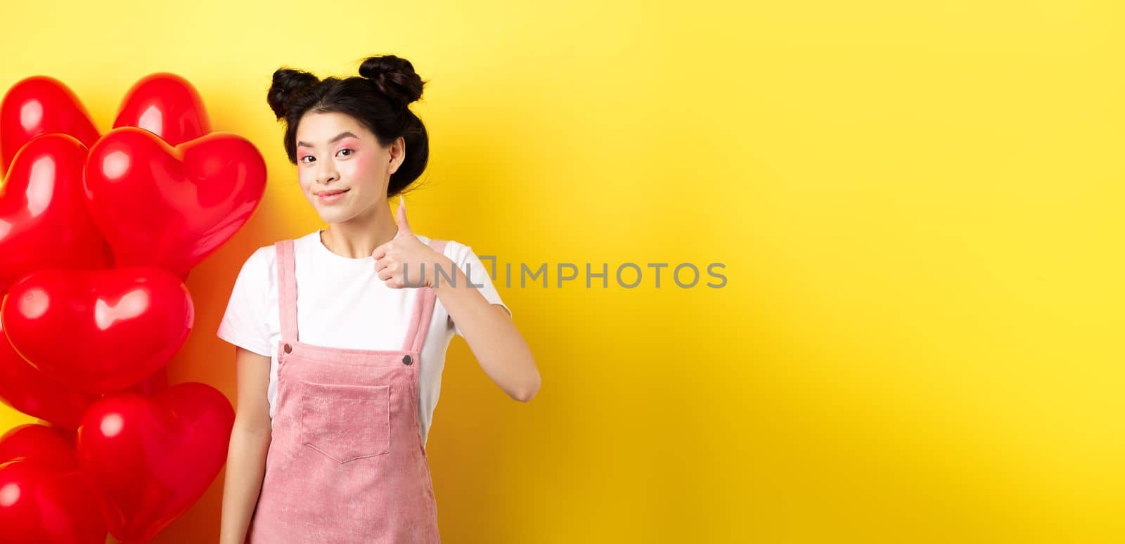 Fashionble asian woman in romantic outfit with make-up, showing thumb up and smiling, praising Valentines day offer, standing near red heart balloons, yellow background.