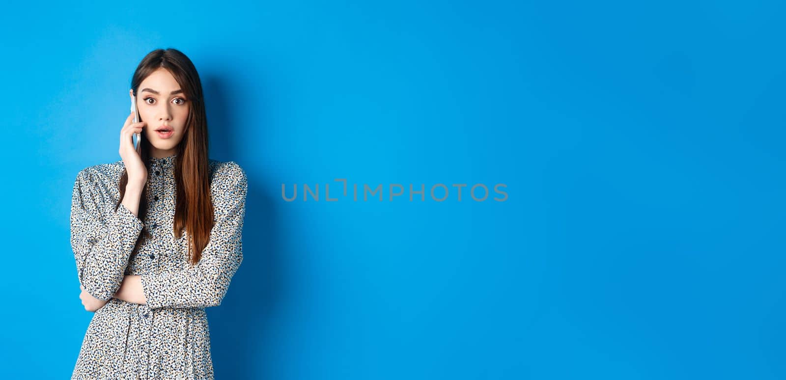 Shocked girl talking on mobile phone, gasping amazed at camera, speaking with someone and hear shocking news, standing in dress on blue background by Benzoix