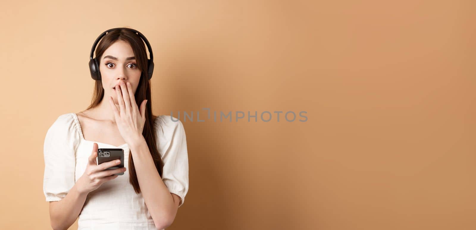 Surprised young woman gasping and covering mouth with hand, using wireless headphones to listen podcast or music, holding mobile phone, beige background.