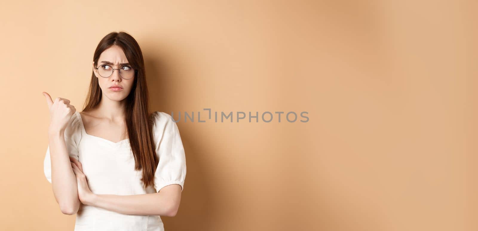 Suspicious woman in glasses pointing and looking aside, frowning with doubtful face, standing on beige background by Benzoix