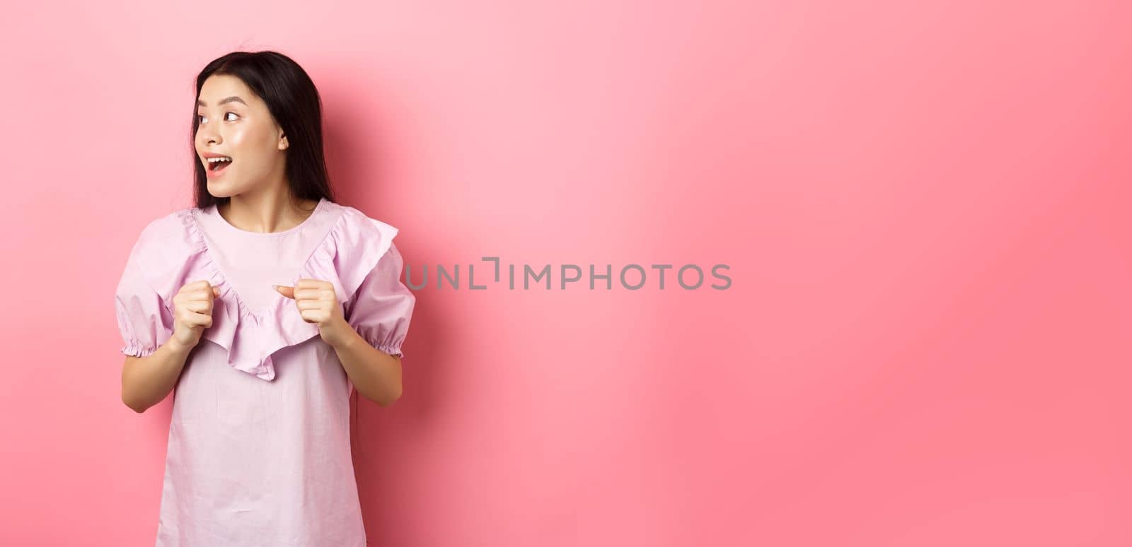 Excited asian girl look left with motivated face, smiling happy, standing in dress on pink background.