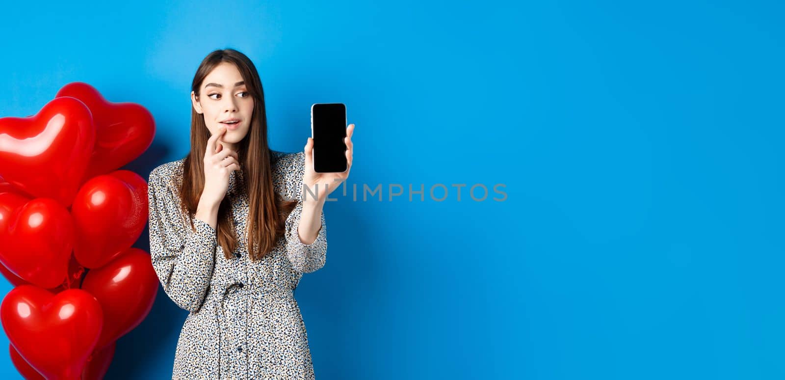 Valentines concept. Beautiful girl looking and showing mobile screen, checking out online shopping offer on lovers day, standing on blue background.