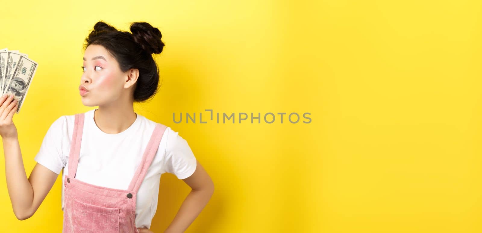 Shopping. Silly korean girl with glam makeup, pucker lips and looking at dollar bills, wasting money, standing on yellow background by Benzoix