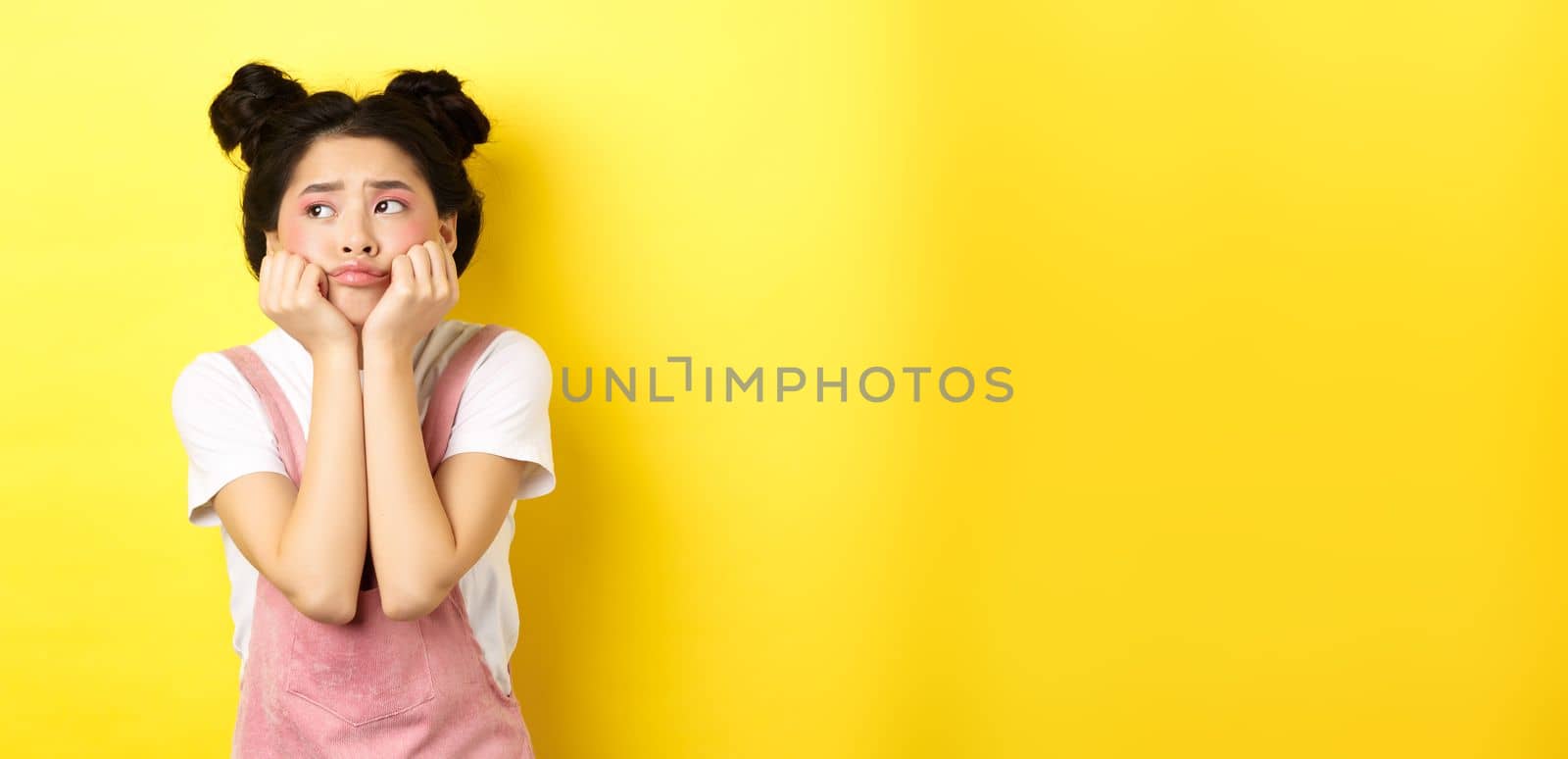 Sad moody asian girl, lean face on hands, looking left with sulking expression, standing in summer clothes on yellow background by Benzoix