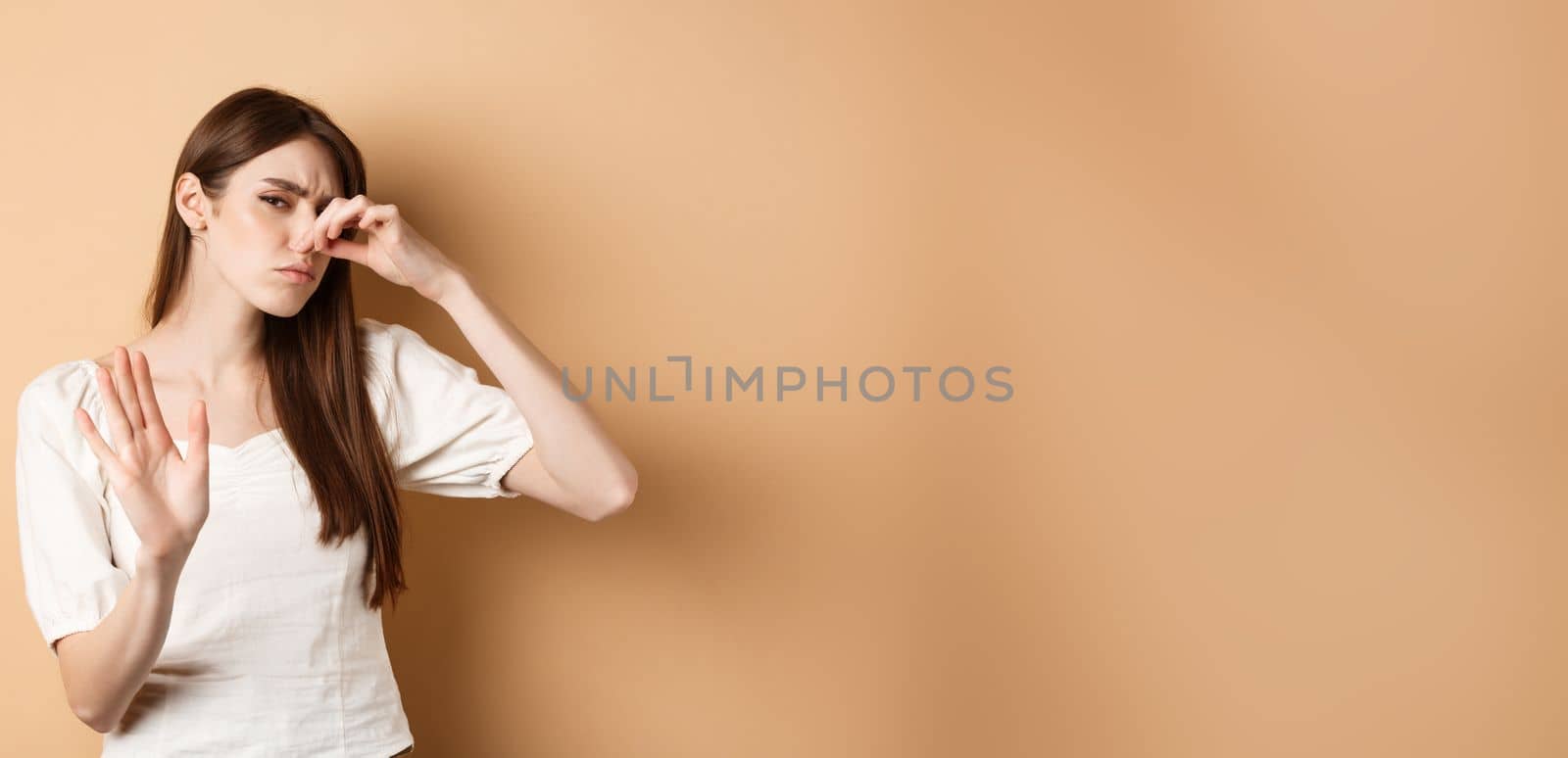 Displeased woman plug her nose from bad smell, showing stop gesture and frowning from disgust. Shut nostrils in aversion, standing on beige background.
