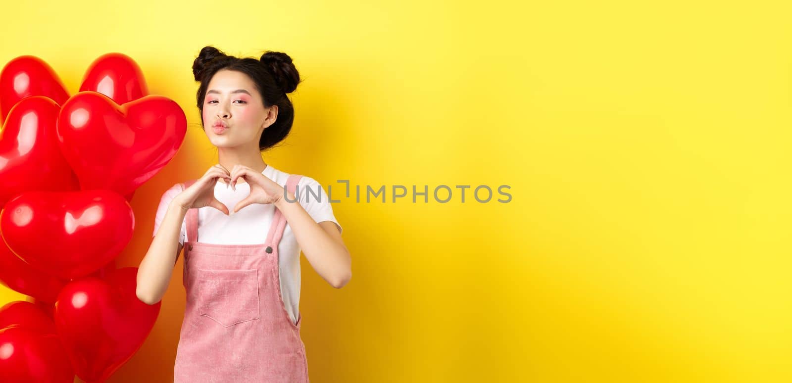 Beautiful asian woman showing heart, I love you gesture and kissing lips, standing near romantic red balloons. Concept of Valentines day.