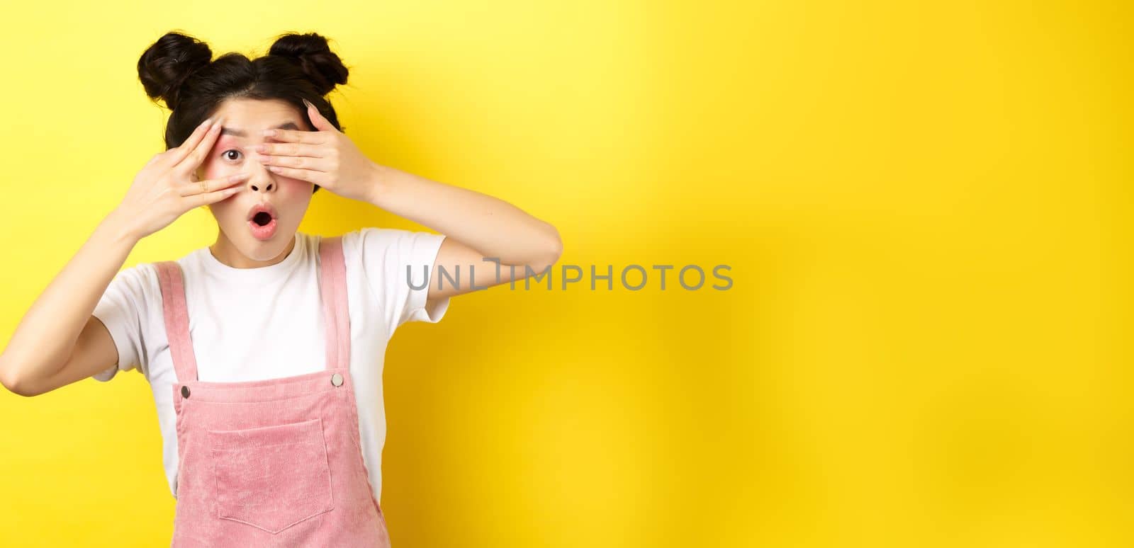 Excited asian teen girl covering eyes with hands and peeking through fingers, gasping amazed at camera, standing in summer clothes on yellow background.