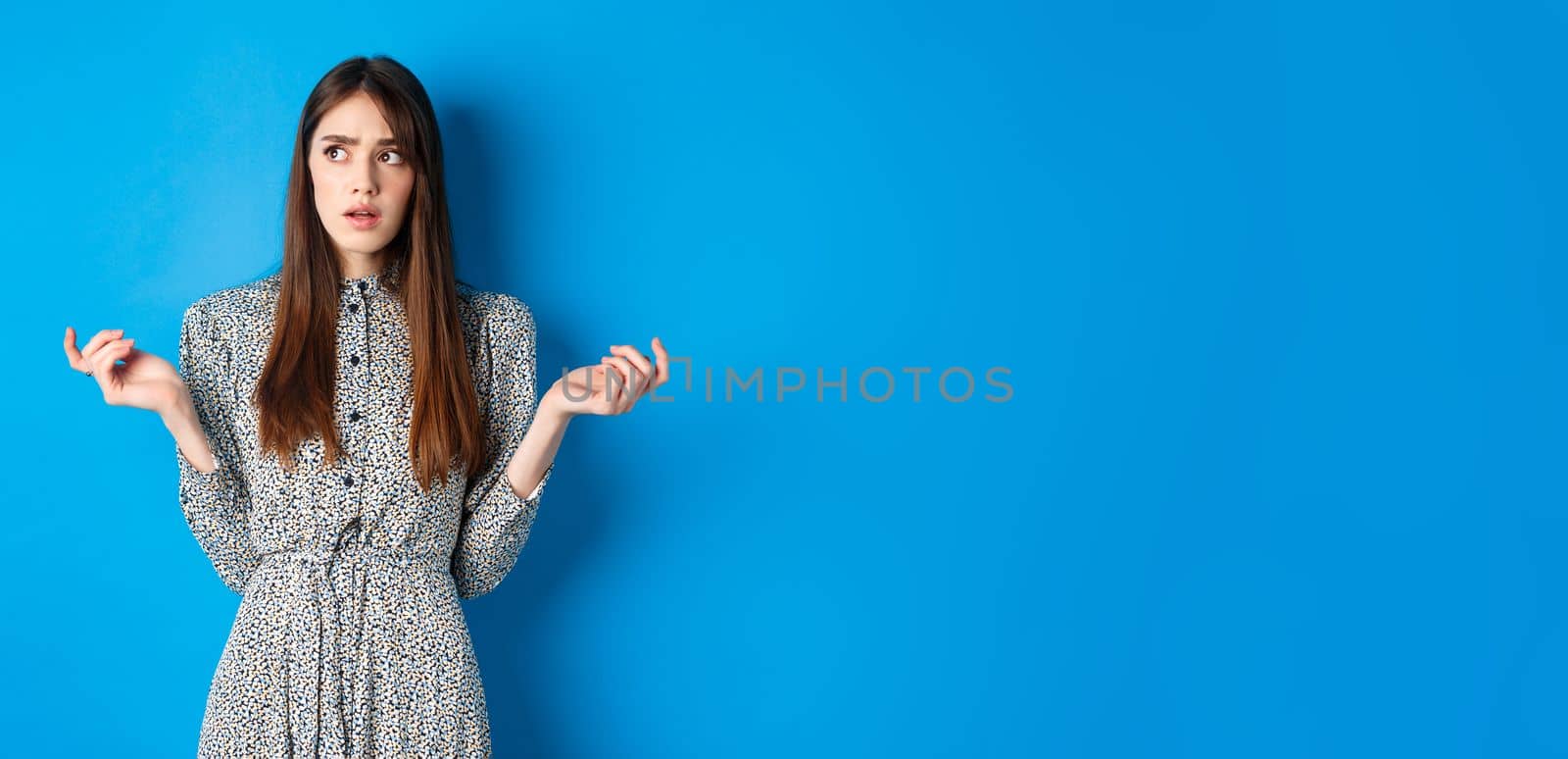 Confused cute girl cant understand something, look away pensive and counting on fingers, standing in dress against blue background.