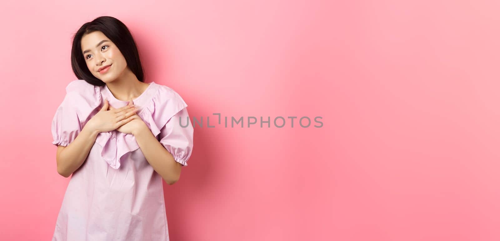 Dreamy asian girl smiling and holding hands on chest, looking left at something romantic and cute, standing on pink background by Benzoix