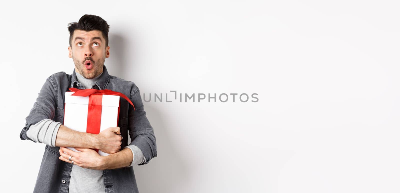 Surprised funny guy hugging big holiday gift and looking up, say wow amazed, checking out Valentines day special offer, standing on white background.