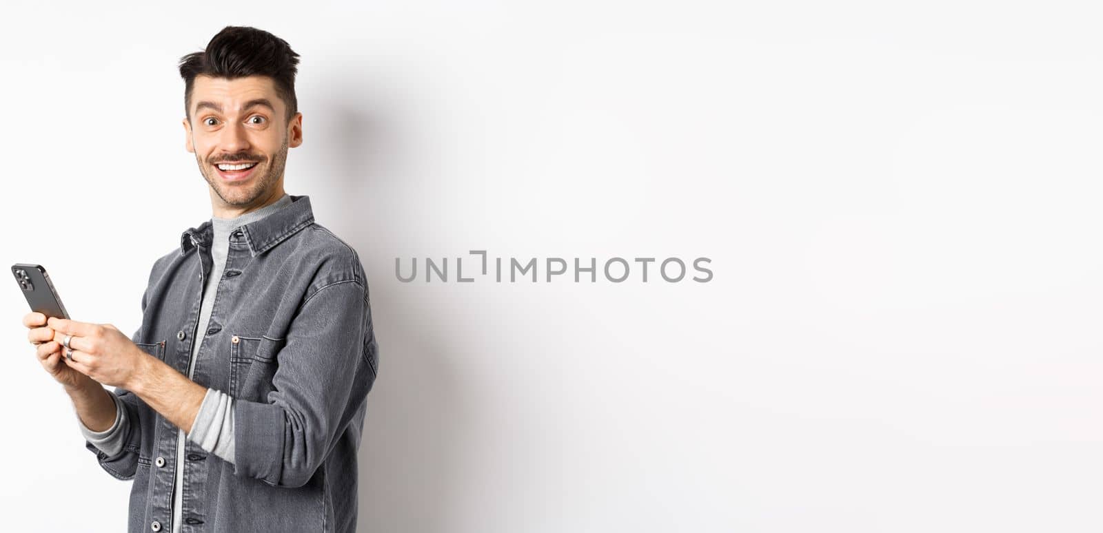 Handsome young man using smartphone and looking at camera. Caucasian guy chatting on phone with happy face, standing on white background.