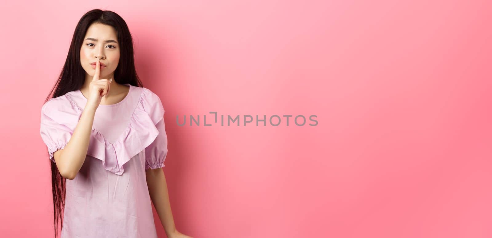 Cute asian girl tell to be quiet, scolding loud person, showing shush sign with finger pressed to lips, standing in dress on pink background.